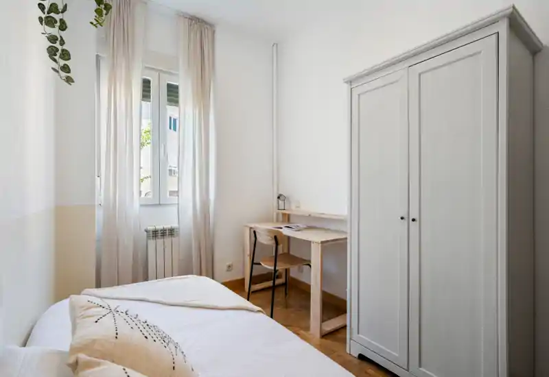 Bright, minimal bedroom with a single bed, light curtains and a small desk next to a wardrobe; neutral tones and natural light make it inviting for a short-term stay.