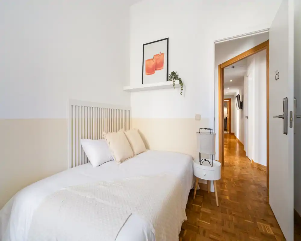 Chambre lumineuse et minimaliste avec un lit simple, linge de lit neutre et coussins doux ; parquet et petite table de chevet créent un coin nuit chaleureux et compact.