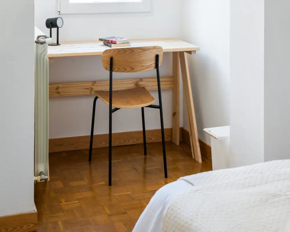 Coin de chambre lumineux et minimaliste avec un bureau simple et une chaise en bois, à côté d'un lit partiellement visible. Espace soigné, parquet chaleureux et lumière naturelle venant de la fenêtre.