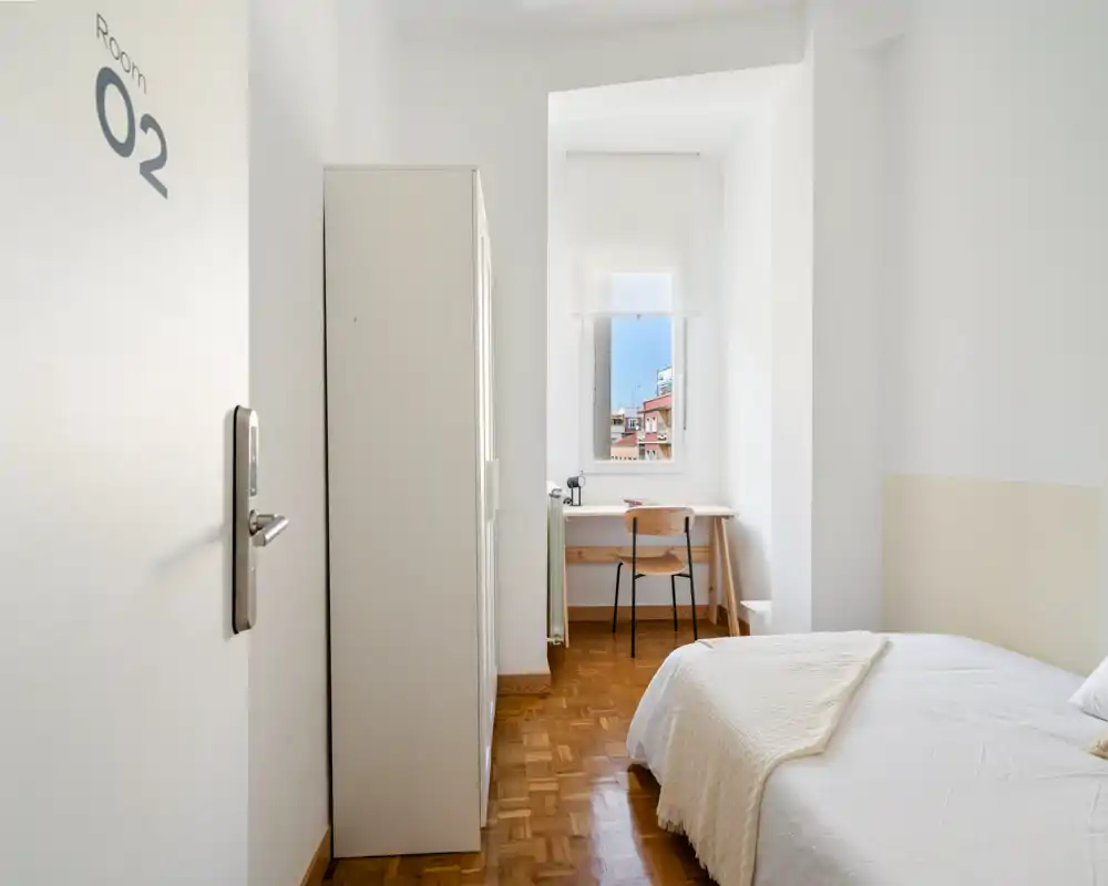 Chambre simple lumineuse et minimaliste avec lit bien fait, armoire et bureau près de la fenêtre offrant une vue sur la ville — idéale pour un court séjour.