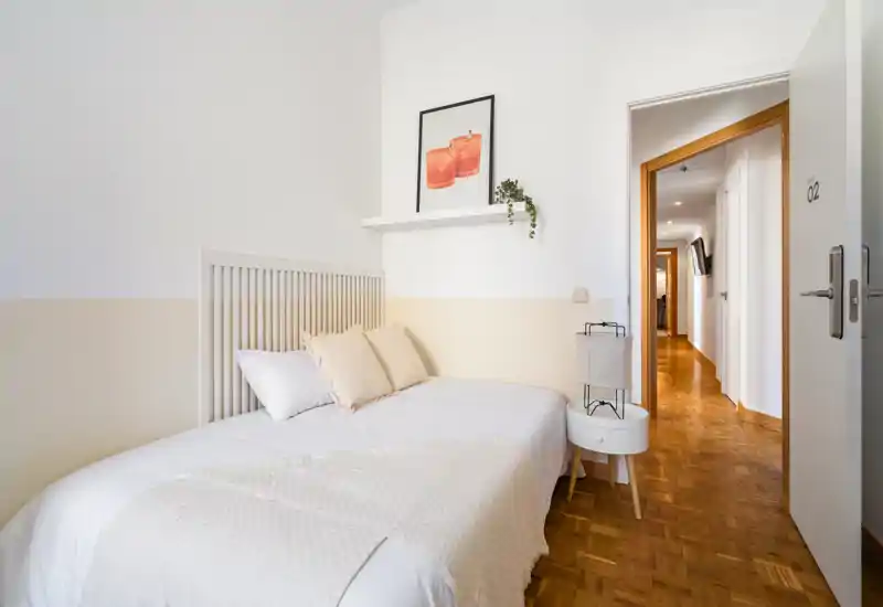 Bright, minimal bedroom with a single bed, neutral linens and soft decorative pillows; clean parquet flooring and a small bedside table create a welcoming, compact sleeping area.