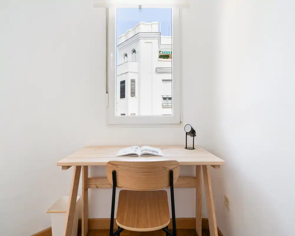 Coin bureau minimaliste et lumineux avec un bureau et une chaise en bois sous la fenêtre, parfait pour étudier ou travailler à distance. Lampe et livre ouvert pour une ambiance épurée.