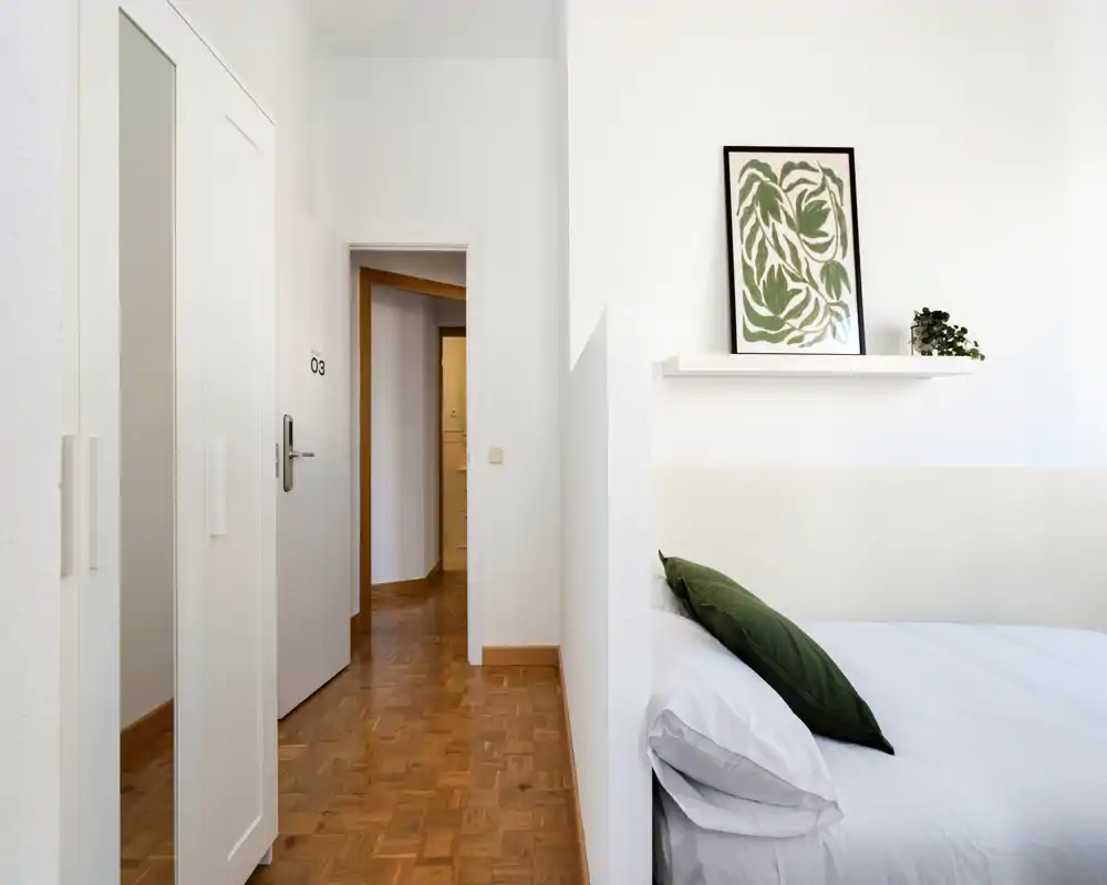 Coin de chambre lumineux et minimaliste avec un lit bien fait, un coussin vert en accent et une étagère murale avec cadre — idéal pour des annonces de location soulignant la déco calme et la lumière naturelle.