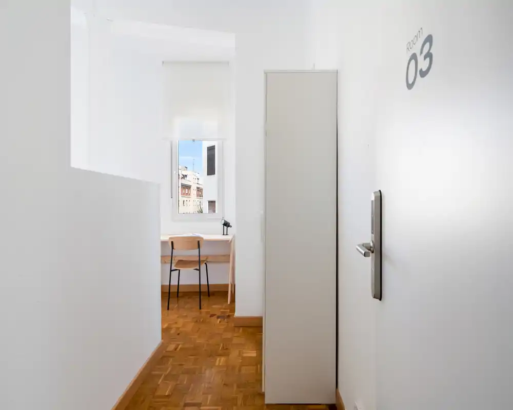Couloir minimal menant à la chambre avec un petit bureau près de la fenêtre ; murs blancs, parquet et une armoire simple créent un espace lumineux et fonctionnel.