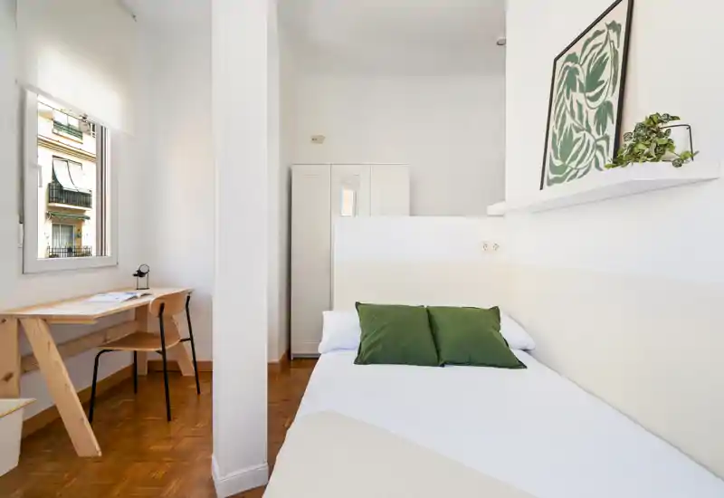 Chambre compacte et lumineuse avec un lit simple en linge blanc et coussins verts, un bureau près de la fenêtre et une armoire à l’arrière — idéale pour étudiant ou séjour solo.