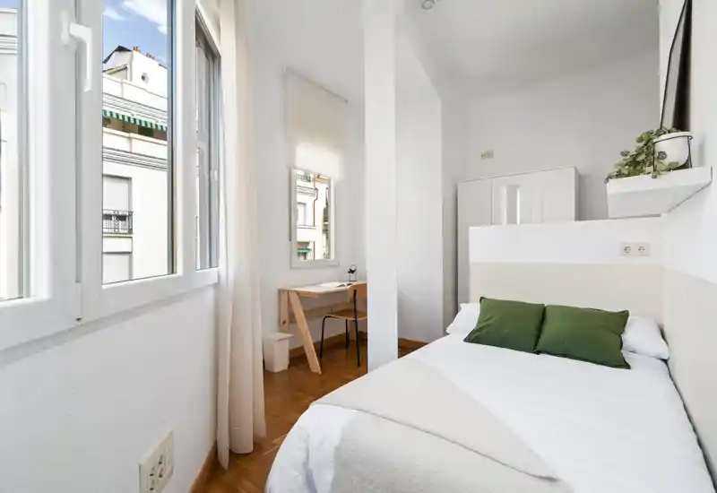 Chambre compacte et lumineuse avec un lit confortable, coussins verts et lumière naturelle d'une grande fenêtre ; bureau et armoire simples pour un espace pratique et aéré.