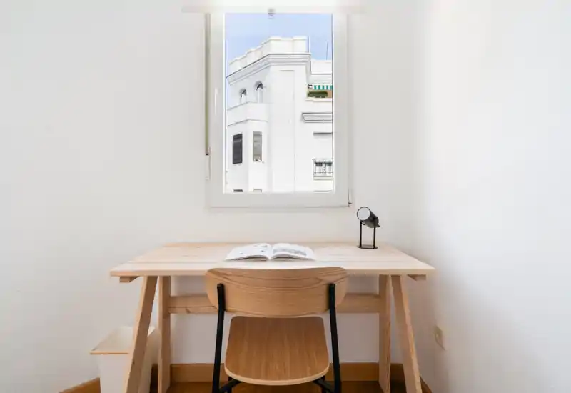 Pequeño espacio de trabajo luminoso con escritorio y silla de madera bajo la ventana, ideal para estudiar o teletrabajar. Lámpara y libro abierto crean una sensación limpia y funcional.