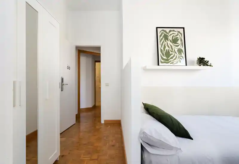 Coin de chambre lumineux et minimaliste avec un lit bien fait, un coussin vert en accent et une étagère murale avec cadre — idéal pour des annonces de location soulignant la déco calme et la lumière naturelle.