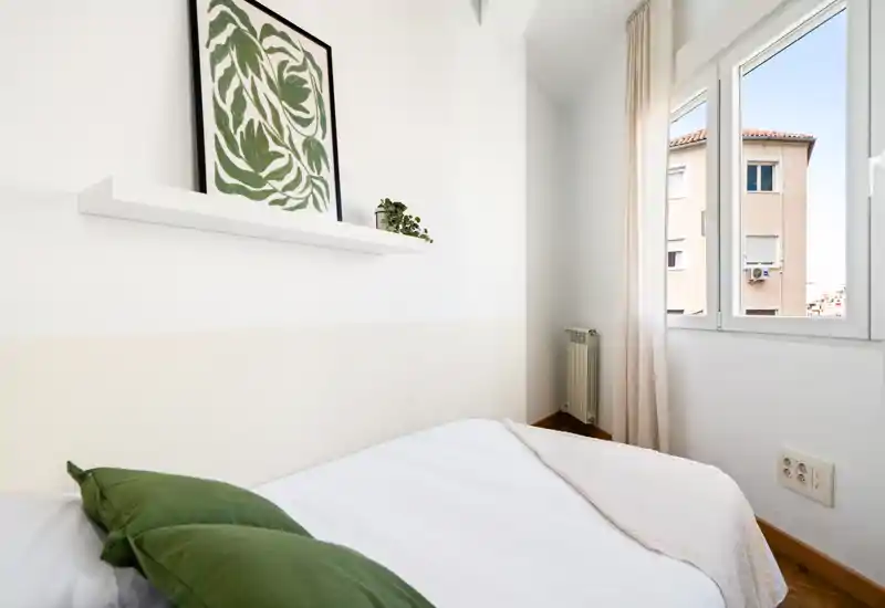Chambre compacte et lumineuse avec lit bien fait, coussins verts et grande fenêtre laissant entrer la lumière naturelle. Décoration minimaliste et petite étagère avec tableau pour une ambiance apaisante.