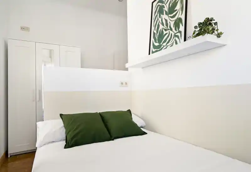 Chambre lumineuse et minimaliste avec un lit bien fait, coussins verts en accent, une armoire blanche et une affiche botanique pour une ambiance fraîche et moderne.