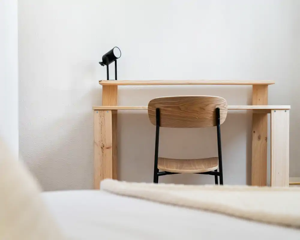 Angolo di camera minimal con scrivania e sedia in legno visibili dietro il bordo del letto; immagine luminosa e nitida con toni caldi in stile scandinavo.