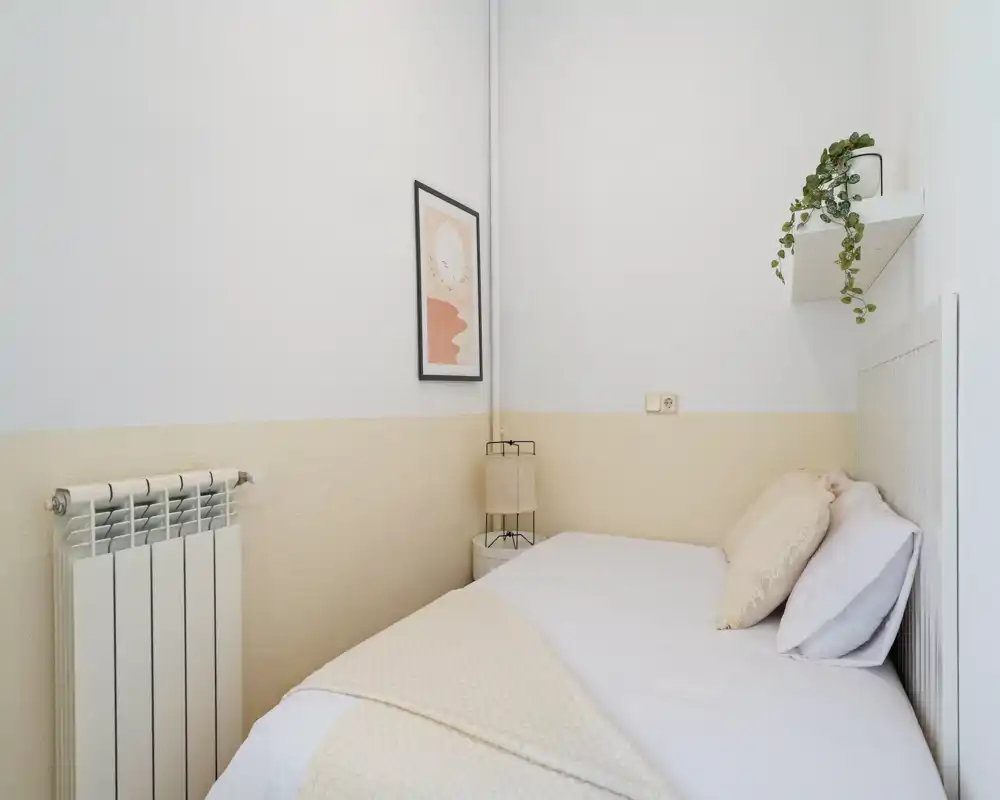 Camera da letto piccola e curata con letto singolo ben rifatto, biancheria neutra e morbida, lampada da comodino e mensola con pianta: luminosa, minimal e accogliente.
