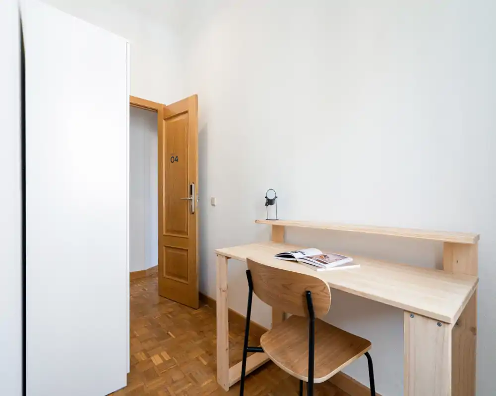 Angolo di camera compatto con scrivania e sedia in legno chiaro accanto a un armadio e una porta aperta, luminoso e arredato in modo semplice — ideale come angolo studio in una stanza in affitto.