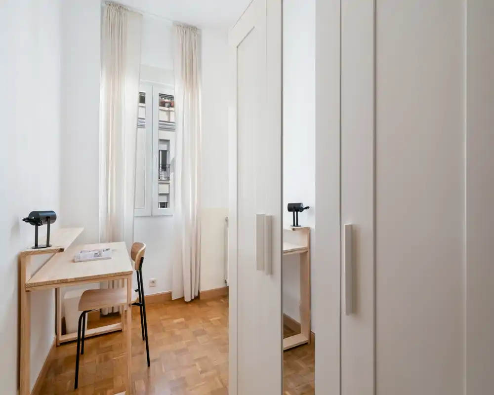 Stanza compatta con un armadio ampio e una semplice scrivania in legno accanto a una finestra luminosa; toni neutri e pavimento in parquet creano un'atmosfera minimal e calma.