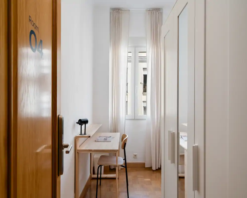 Camera singola compatta con una piccola scrivania in legno, sedia e armadio; luce naturale morbida dalle tende velate rende l'ambiente luminoso e tranquillo.
