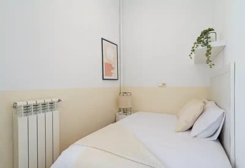 Small, stylish bedroom with a neatly made single bed, soft neutral linens, a bedside lamp and a wall shelf with plant—bright, minimal and cozy.