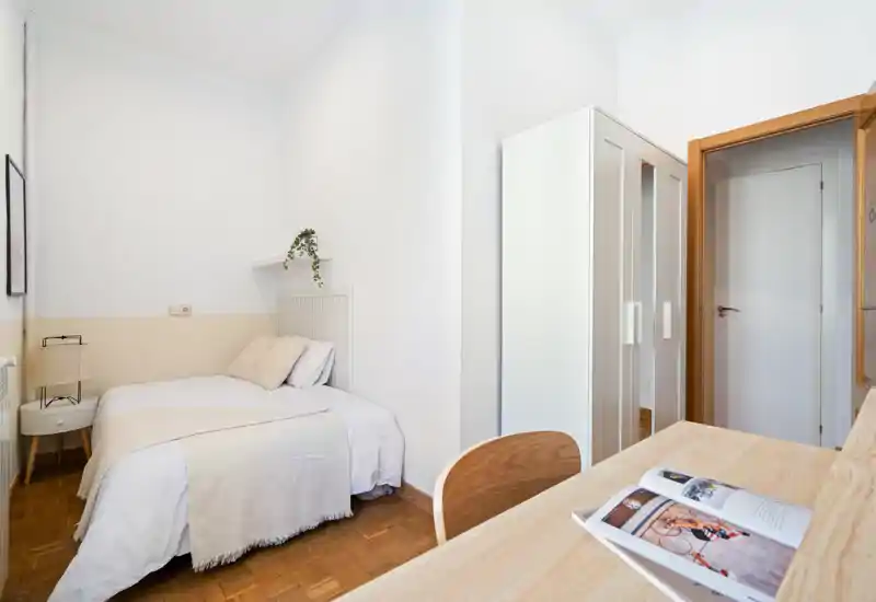 Bright, minimal bedroom with a neatly made double bed, bedside table and lamp, wardrobe and a small desk in the foreground. Neutral tones and natural light create a calm, welcoming atmosphere ideal for rental listings.
