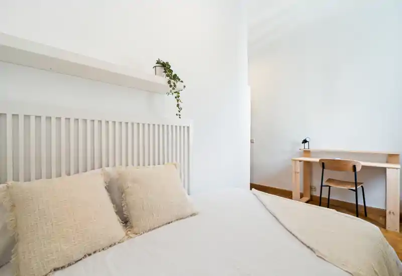 Bright, minimal bedroom with a large bed dressed in neutral linens, soft textured pillows, and a small wooden desk in the corner — a calm, airy space ideal for restful stays.