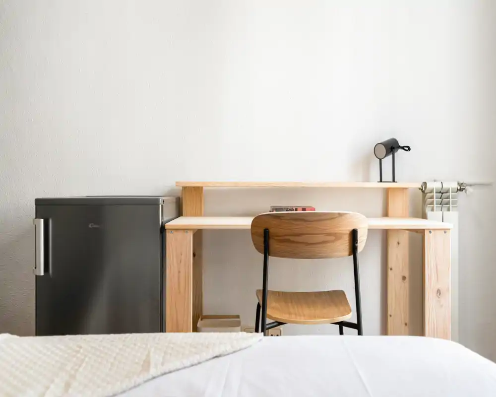 Minimal, bright bedroom corner with a wooden desk and chair beside a compact fridge; crisp bedding is partially visible in the foreground, ideal for a work-and-sleep studio setup.