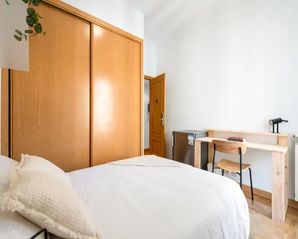 Bright, minimal bedroom with a comfortable double bed, built-in wooden wardrobe and a simple desk — ideal for a cozy single or couple stay.
