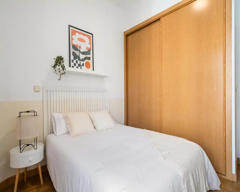 Bright, neatly styled bedroom with a double bed, bedside table and built-in wooden wardrobe—calm neutral tones and subtle decorative details for a welcoming stay.