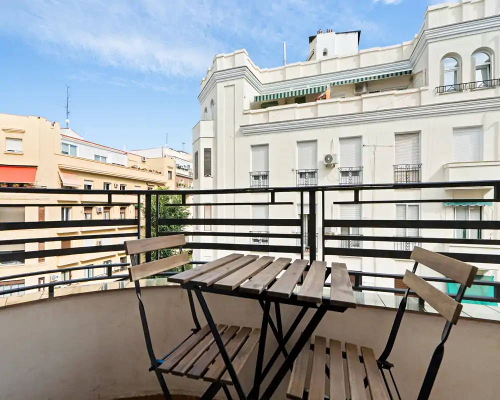 Sunny private balcony with a small folding wooden table and chairs, overlooking neighboring residential buildings — ideal for morning coffee.