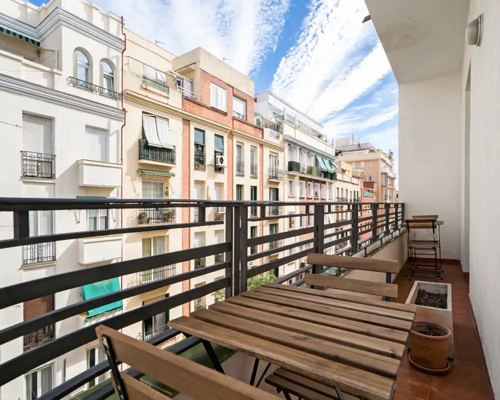 Sunny narrow balcony with wooden bistro table and chairs overlooking neighboring apartment façades — bright urban outdoor space ideal for morning coffee.
