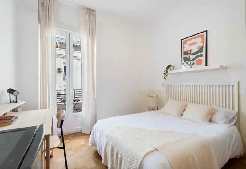 Bright, minimal bedroom with a double bed, soft neutral textiles, a desk by the window and a small balcony access — clean, airy and well-styled for rental photos.