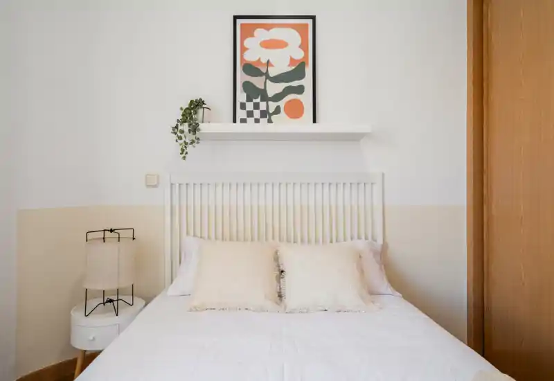 Bright, well-styled bedroom with a white slatted headboard, soft neutral bedding, decorative pillows and a bedside lamp — a cozy, minimal space ideal for restful stays.