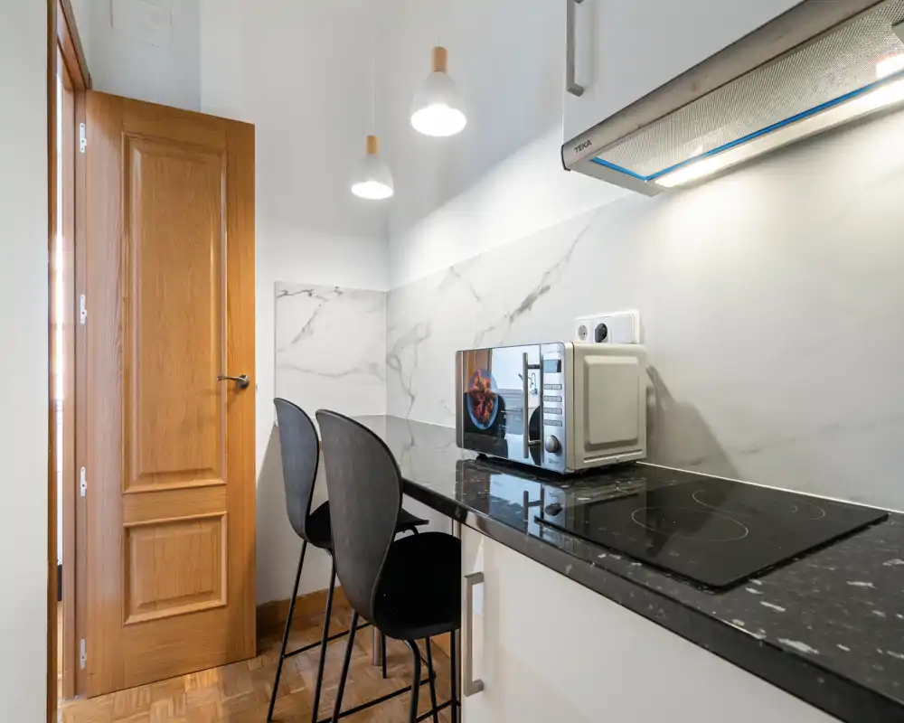 Compact modern kitchen with a glossy black countertop, induction hob, microwave and two bar stools — bright, stylish and neatly arranged for convenient cooking or quick meals.
