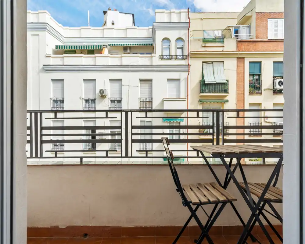 Sunny balcony with a small wooden bistro table and folding chairs, tiled floor and metal railing overlooking neighboring buildings — ideal for morning coffee.