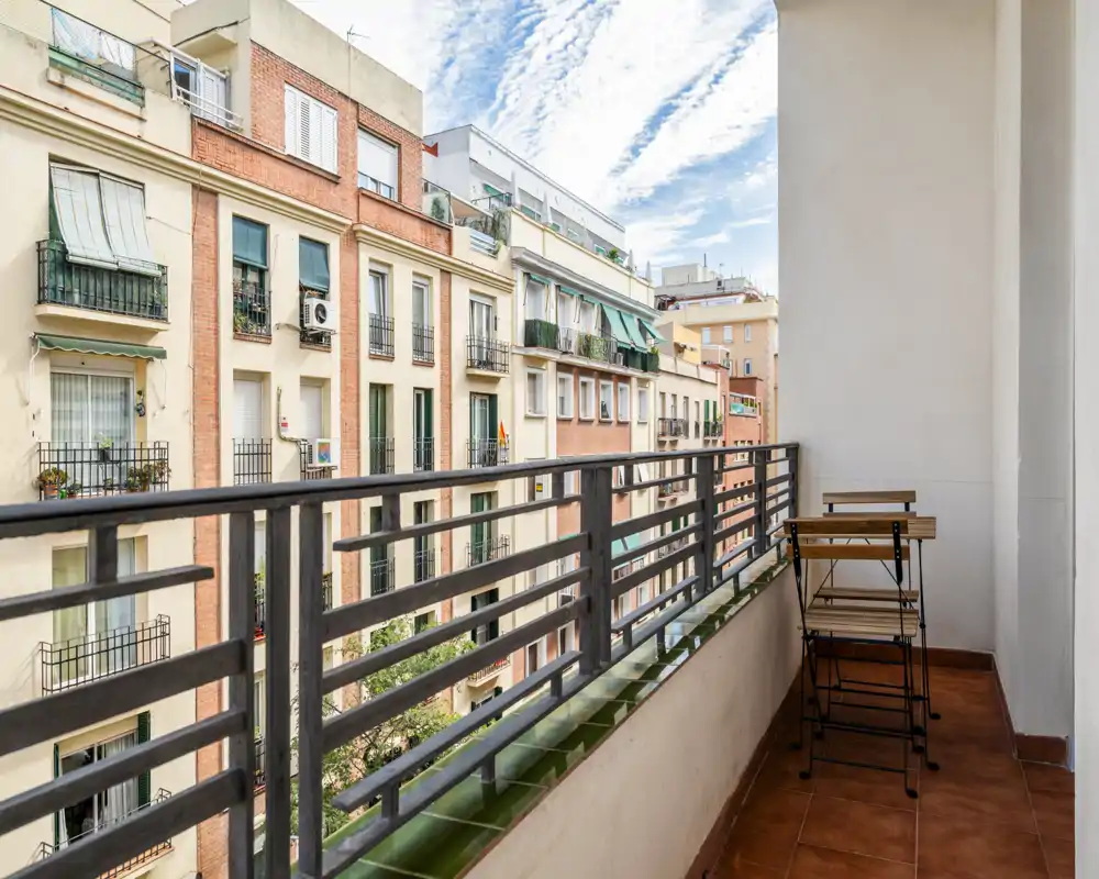 Sunny narrow balcony with a small foldable table and two chairs, overlooking neighboring apartment buildings — ideal for a morning coffee in the city.