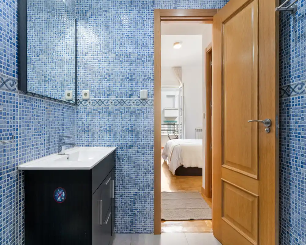 Bright bathroom with blue mosaic tiles, a modern sink vanity and a large mirror; door open to a bedroom with natural light.