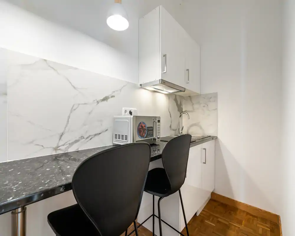 Compact modern kitchenette with a dark marble-effect counter, two black bar stools, microwave and integrated sink — bright and neatly presented for a small apartment.