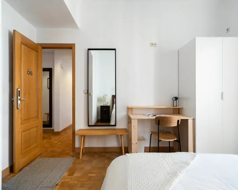 Bright, minimally styled bedroom with warm wood flooring, a visible bed in the foreground, desk and wardrobe along the wall — clean and welcoming for rental listings.