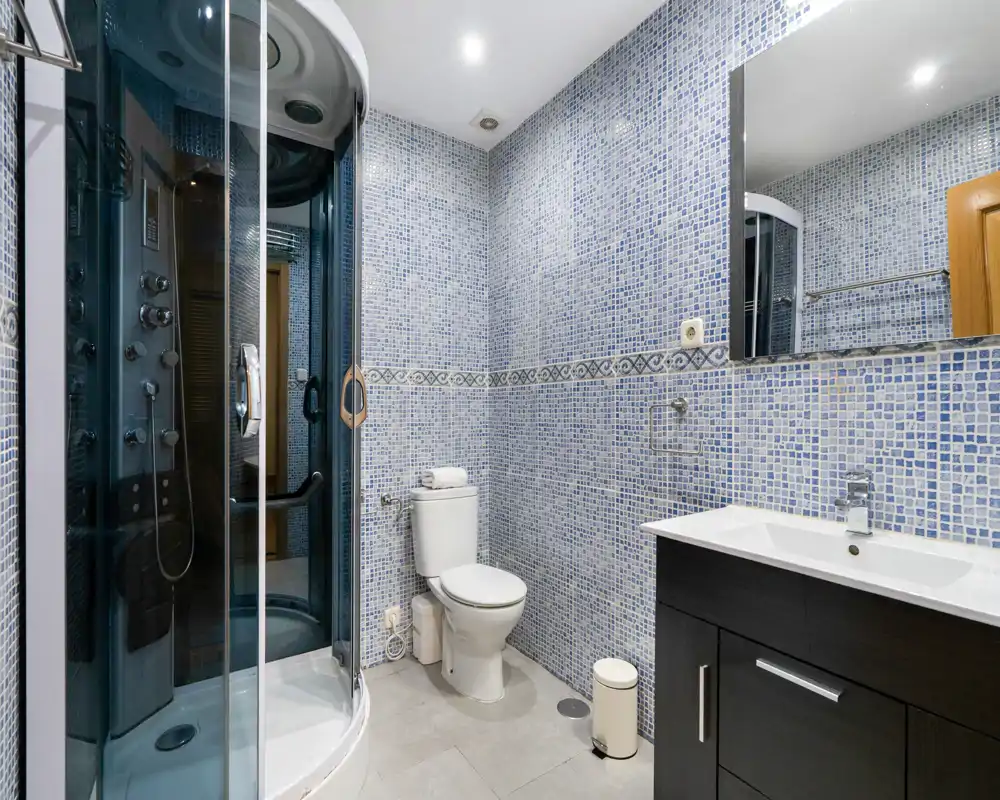 Modern bathroom with mosaic-tiled walls, a glass-enclosed shower cabin, toilet and a dark wood vanity with sink; bright, clean and well-lit.