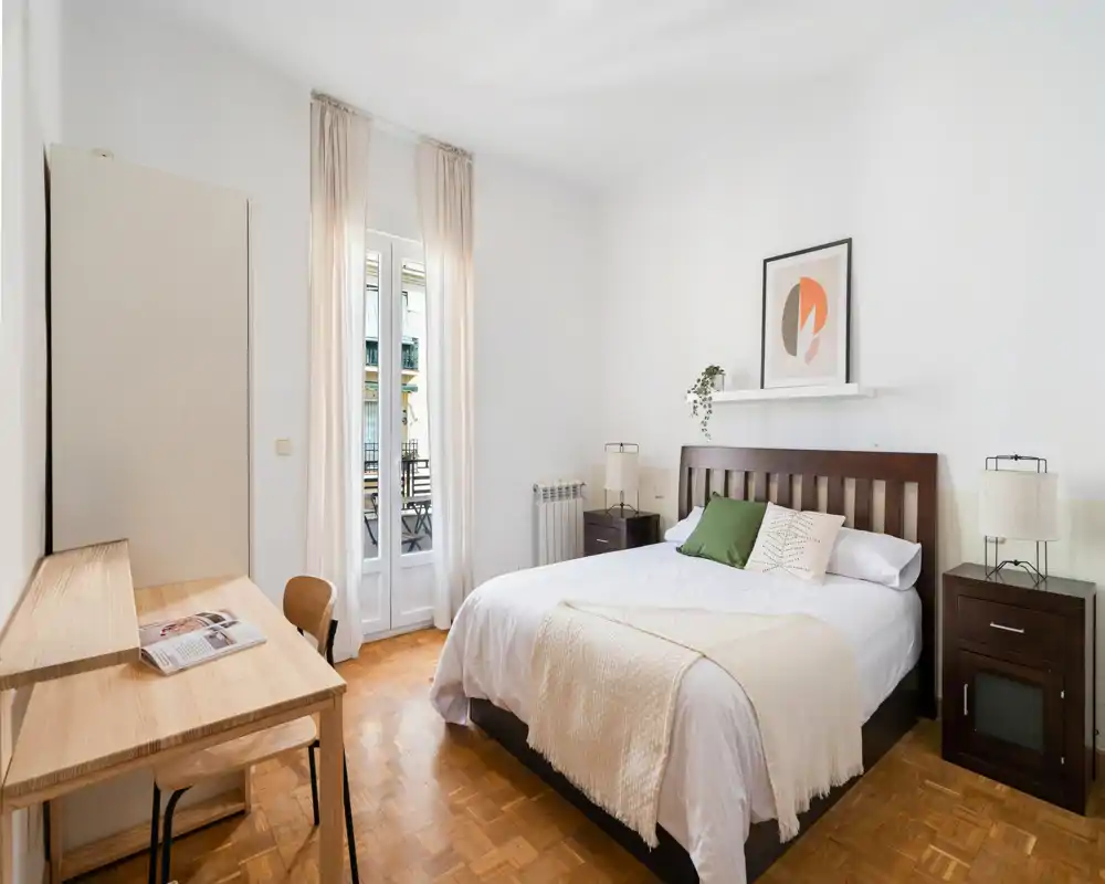 Bright, neatly styled bedroom with a double bed, bedside tables and large window opening to a small balcony; neutral tones and natural light create a calm atmosphere.