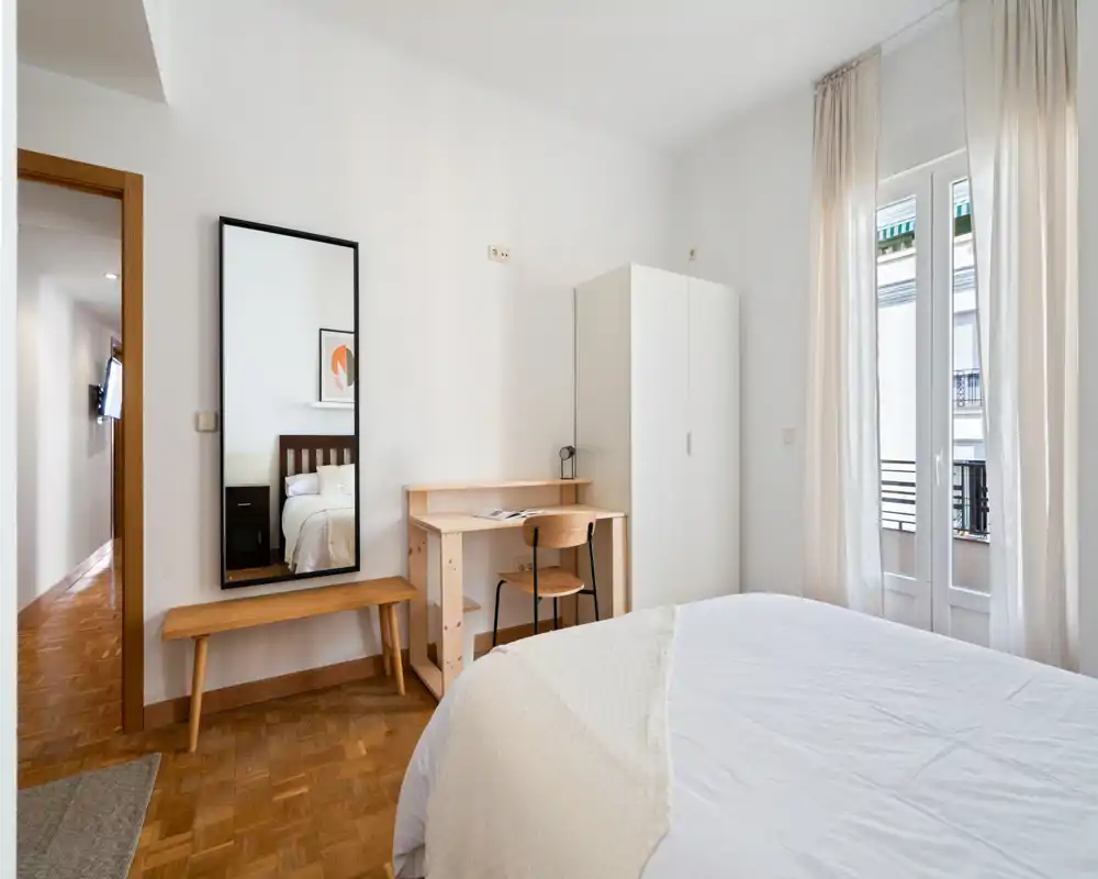 Bright, minimalist bedroom with a comfortable double bed, wooden flooring, a small desk and wardrobe next to a balcony door that fills the room with natural light.