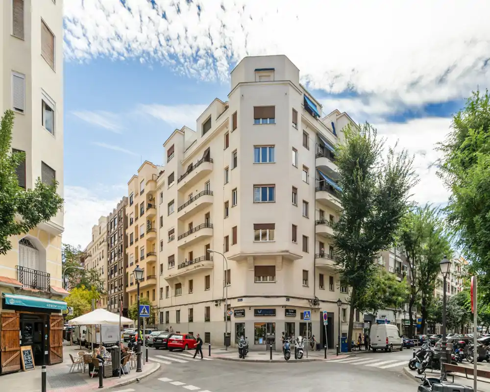 Bright urban street corner with a mid-rise residential building, sidewalk cafe seating and parked vehicles — ideal to show neighborhood character and locale.