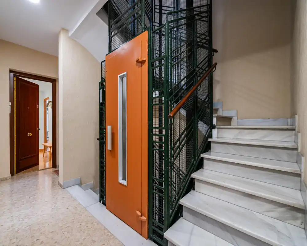 Entrance/stairwell with a vintage-style elevator: a bright hallway showing marble stairs, a metal-caged lift with an orange door, and an open apartment doorway.