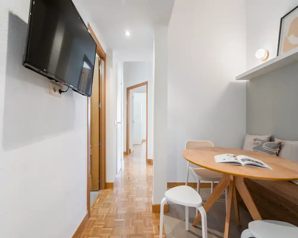 Compact, well-lit living area with a small round dining table, built-in bench with cushions and a wall-mounted TV; bright neutral palette and parquet floor create a cozy, modern feel.