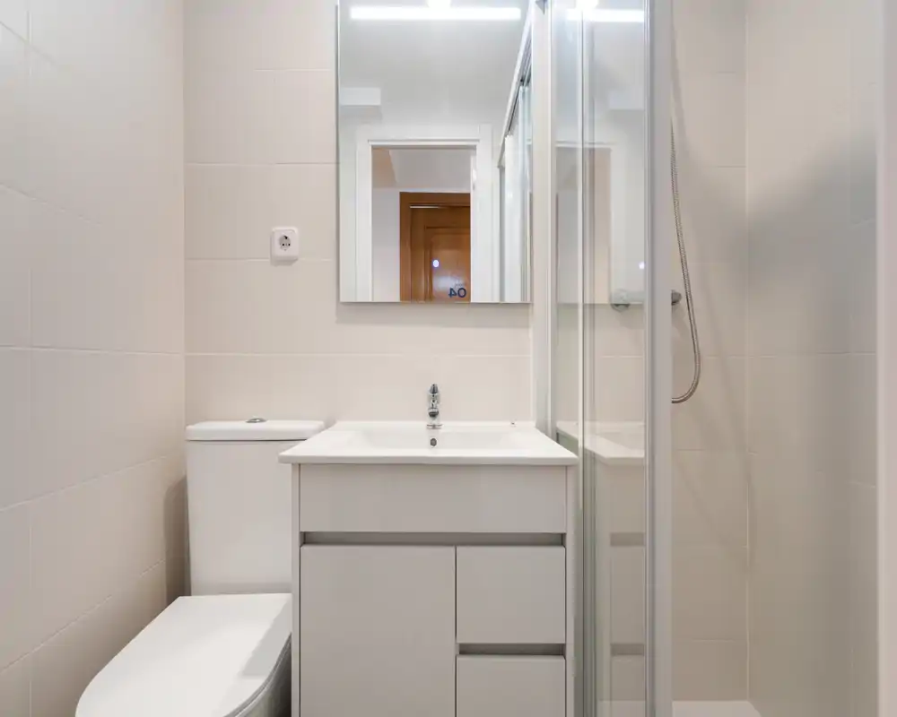 Bright, modern bathroom with a sink vanity, mirror, toilet and a glass-enclosed shower. Clean white tiles and compact, functional layout ideal for city apartments.