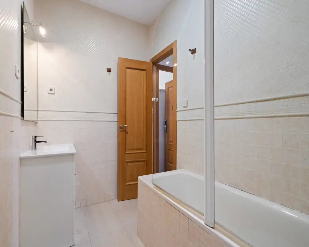 Bright, clean bathroom with a bathtub and sink; neutral tiles and wooden door create a simple, well-lit bathing space.