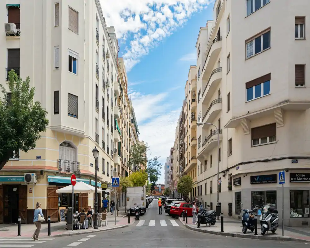 Sunlit urban street with mid-rise residential buildings, parked cars and scooters, a sidewalk café and pedestrians — ideal for showcasing neighborhood and location.