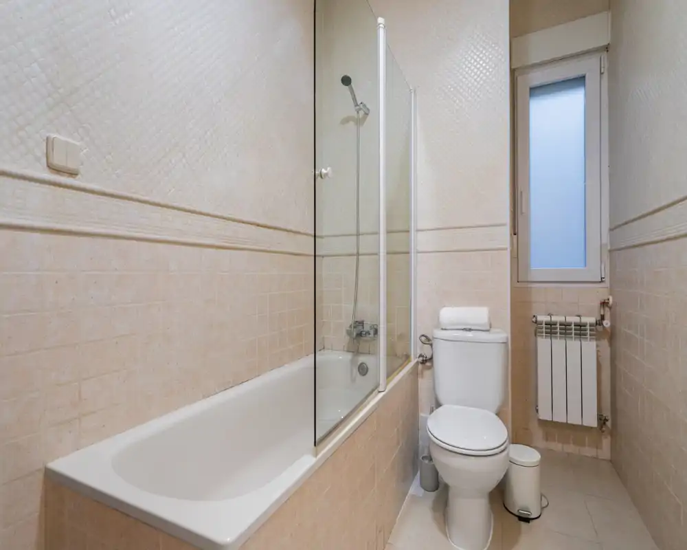 Neat, well-lit full bathroom with a bathtub and shower screen, toilet, and radiator by the window — clean neutral tiles and functional layout.