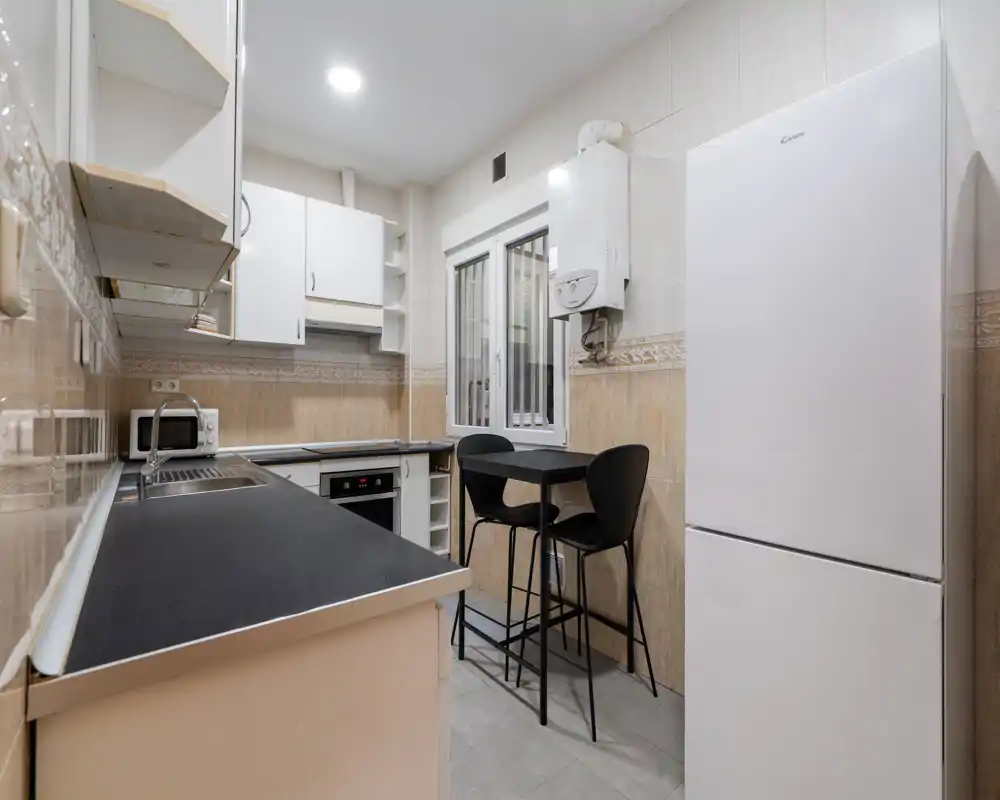 Compact, modern kitchen with black countertops, integrated oven, tall refrigerator and a small high dining table for two — bright and neatly arranged.