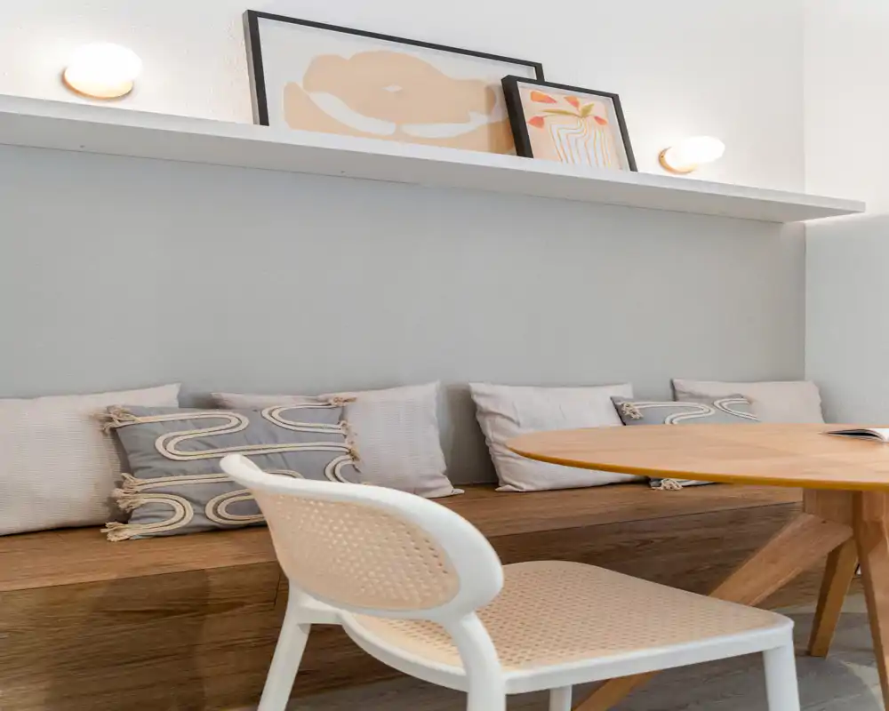 Cozy, modern dining nook with a built-in wooden bench, decorative cushions and a round wooden table—bright, well-styled space ideal for relaxed meals.