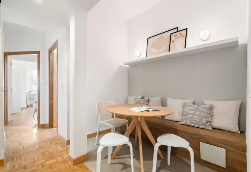 Acogedor rincón comedor luminoso con mesa redonda de madera, banco con cojines y taburetes — ideal para comidas en un espacio compacto.