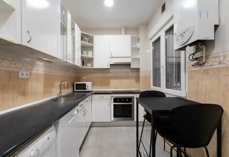 Cocina luminosa y ordenada con muebles blancos, encimeras negras, horno empotrado y una pequeña mesa con sillas — práctica y bien iluminada para el uso diario.
