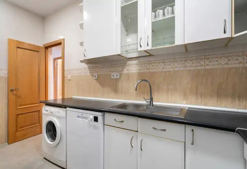 Cocina luminosa y funcional con mobiliario blanco, electrodomésticos integrados y fregadero de acero inoxidable — distribución limpia y práctica para el uso diario.